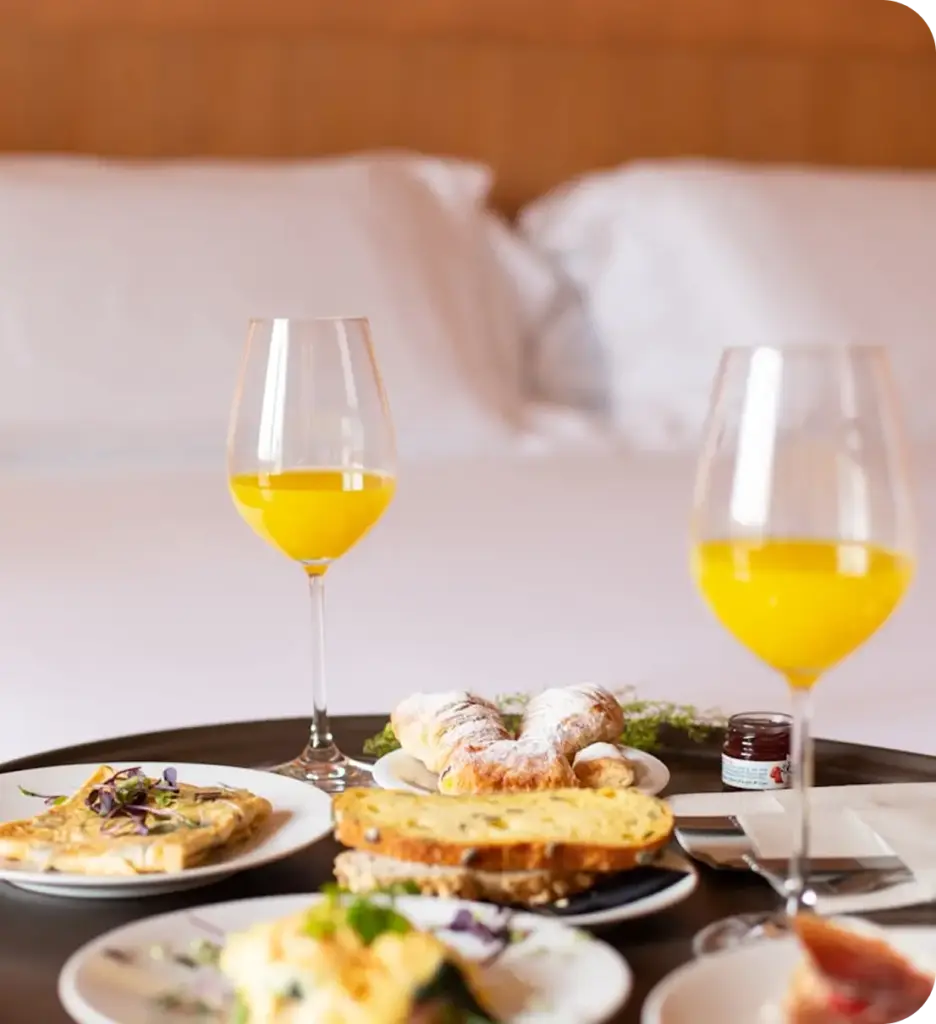 Rahat bir otel odası ortamında, yatakta servis edilen kahvaltı; portakal suyu, hamur işleri ve taze yiyecekler.