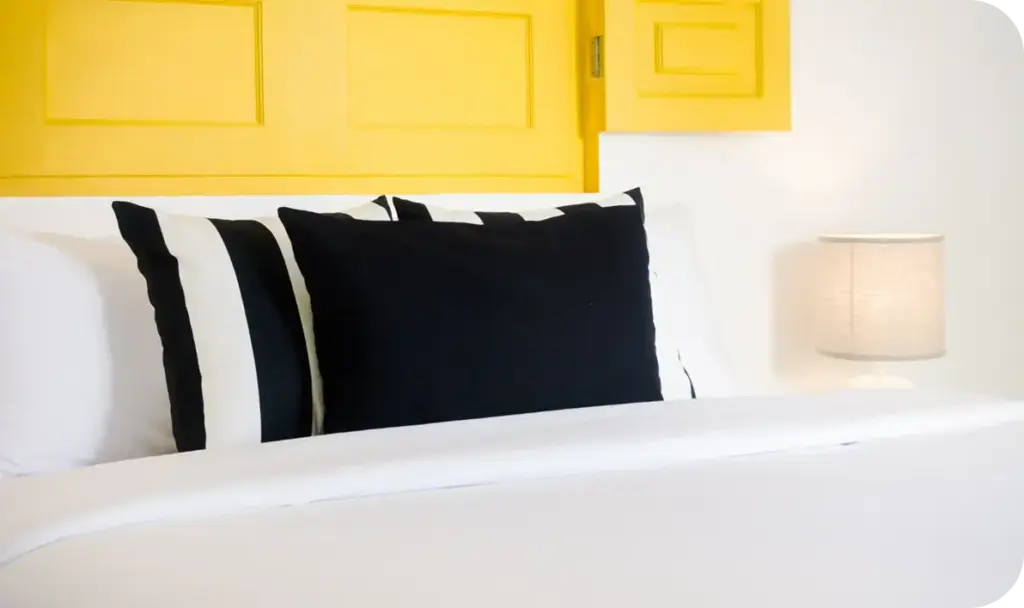 Beyaz yatak, siyah yastıklar ve sarı duvar detaylarıyla şık ve minimalist bir iç mekan yaratan modern yatak odası.