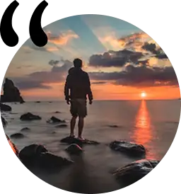 Gün batımı sırasında bir göl kenarında duran kişi; huzurlu bir manzara içinde dağlar ve suyun yansıması.